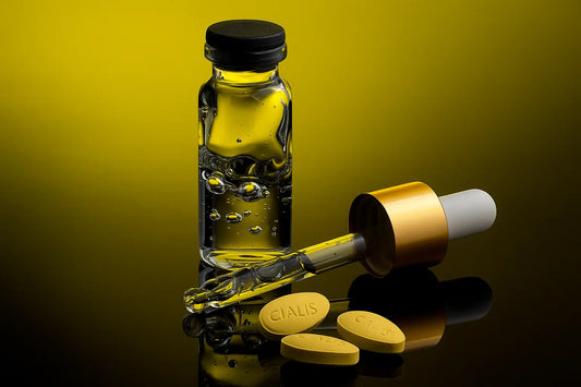 A clear glass vial filled with transparent liquid sits beside a gold-capped dropper and several yellow Cialis tablets on a reflective black surface with a yellow gradient background.